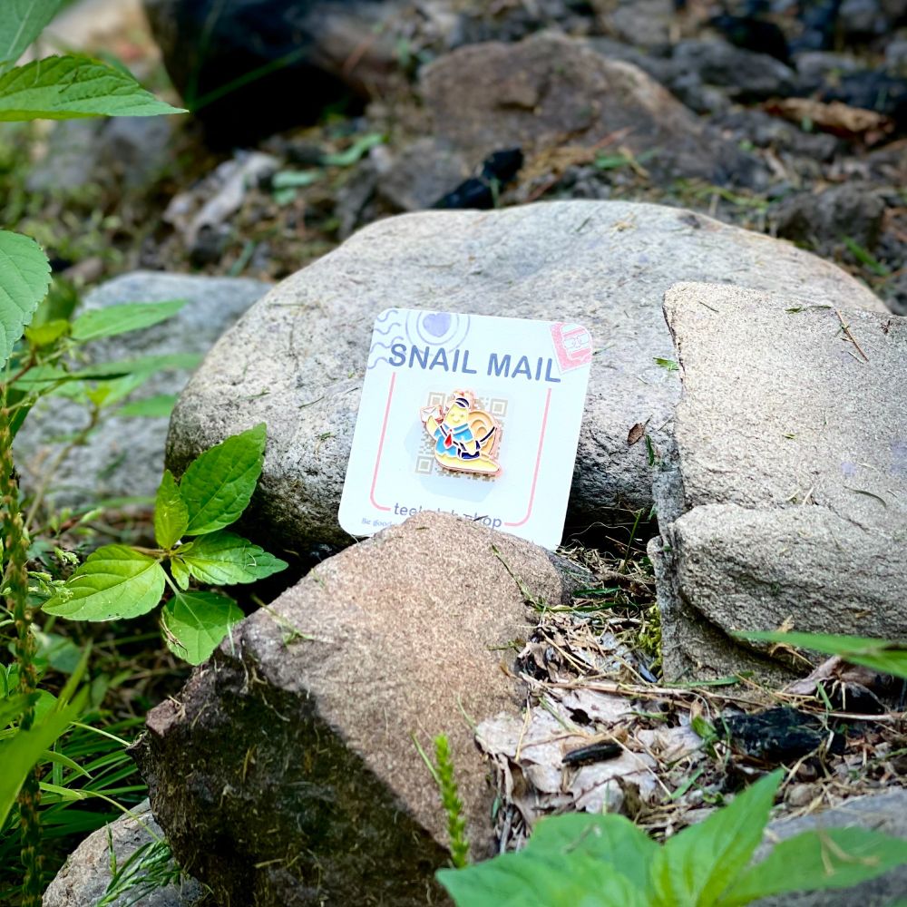 a photo of the snail mail enamel pin on its backing card on some rocks and twigs. The pin is of a small, yellow snail with a mail carriers shirt, tie, cap and bag as they hold up a tiny letter on its way to a friend.