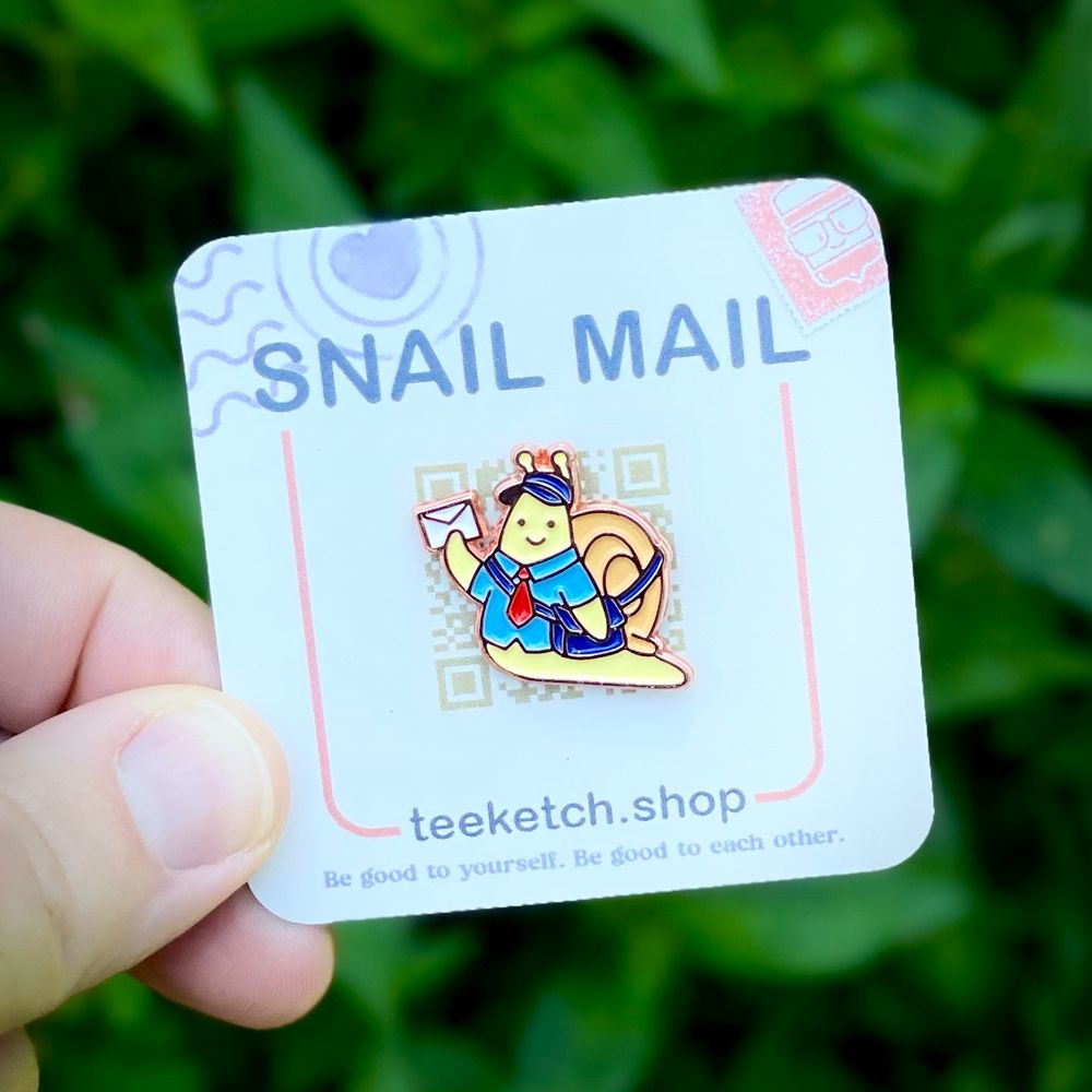 a photo of the snail mail enamel pin on its backing card held in my hand against a background of some plants and shrubs. The pin is of a small, yellow snail with a mail carriers shirt, tie, cap and bag as they hold up a tiny letter on its way to a friend.