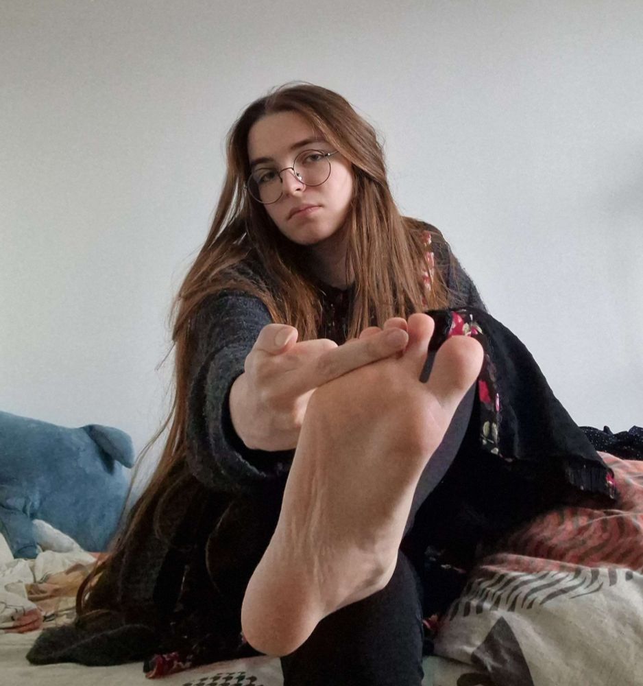 a selfie, i'm sitting down. i'm wearing a dress with a floral pattern on the upper half and a cardigan, you can see i removed the socks that were in the previous pictures. i'm doing a middle finger to the camera by placing said finger against my toes, hiding them a little bit