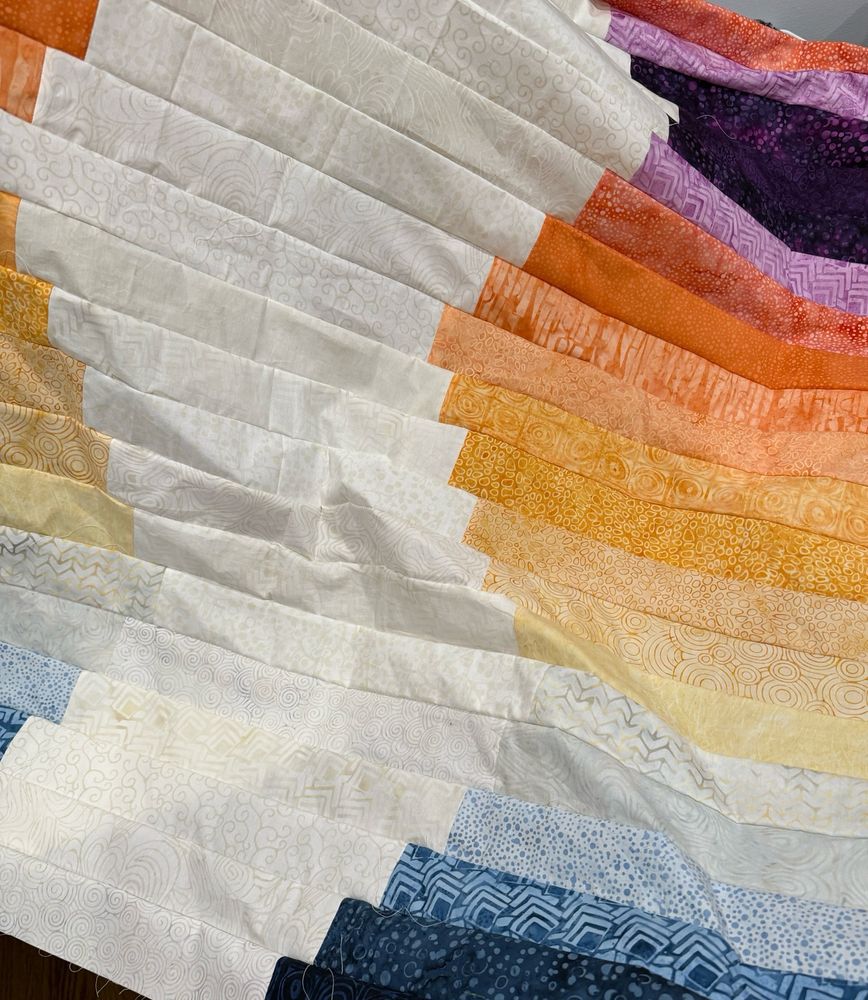 A quilt in progress. Strips of fabric arranged in a gradient (blue to yellow to orange to purple) with a white offset pathway running the length of the quilt.