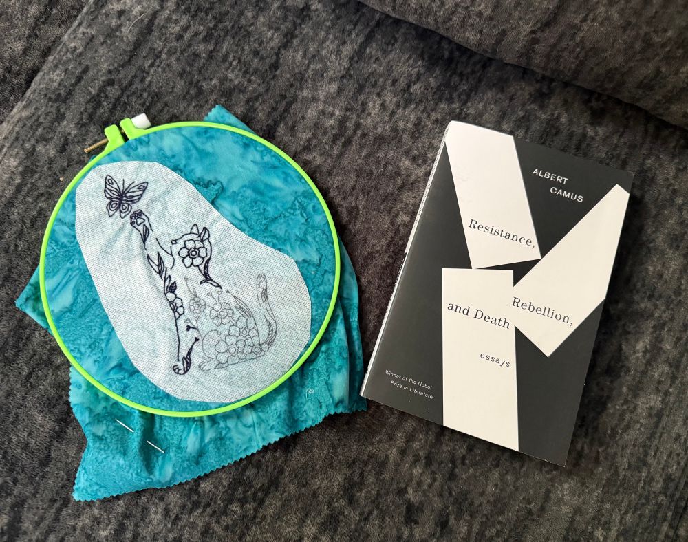 On a gray couch cushion rest a partly-completed embroidery project of a floral cat reaching for a butterfly and a paperback copy of “resistance, rebellion, and death” by Albert Camus.