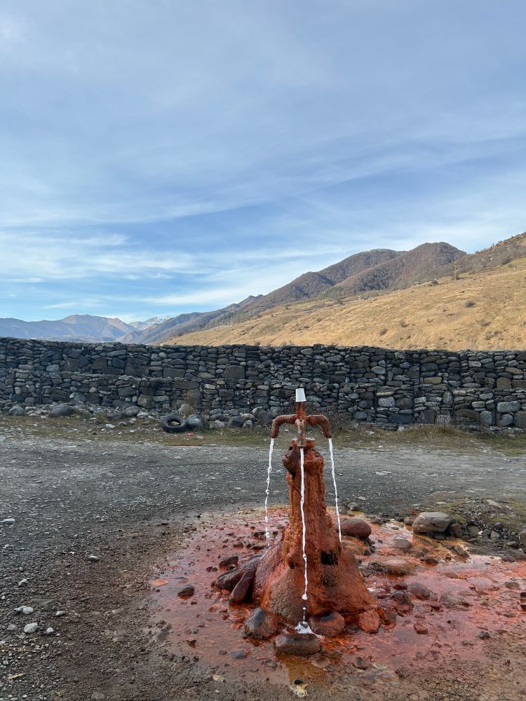 美しい山々をバックに、赤銅色に固まった水道管から天然水が吹き出し流れ落ちている