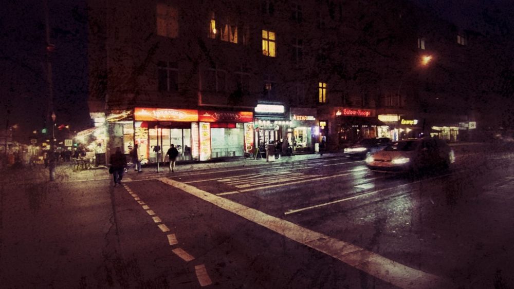 Nachtaufnahme. Im Hintergrund beleuchtete Kneipen und Dönerstände, im Vordergrund eine breite Straße mit einem heran nahenden Auto. Links teilweise ein Gehstreifen sichtbar. Vor der Häuserzeile mit den beleuchteten Schaufenstern und Eingängen sind Menschen umrissartig sichtbar. Beim Foto ist ein Filter gesetzt der Farben und Lichter stark betont