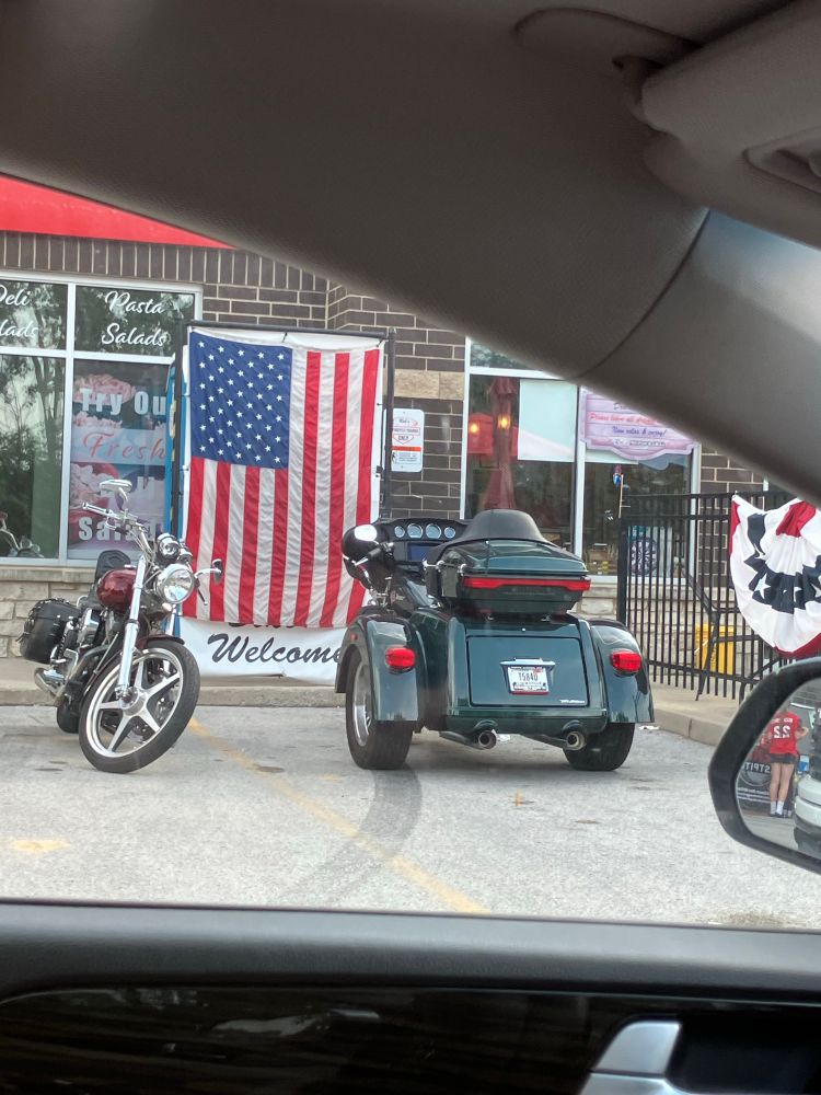 A large American flag with the word “welcome” underneath it. You can see a blue portapotty behind it. There are two motorcycles parked in front.