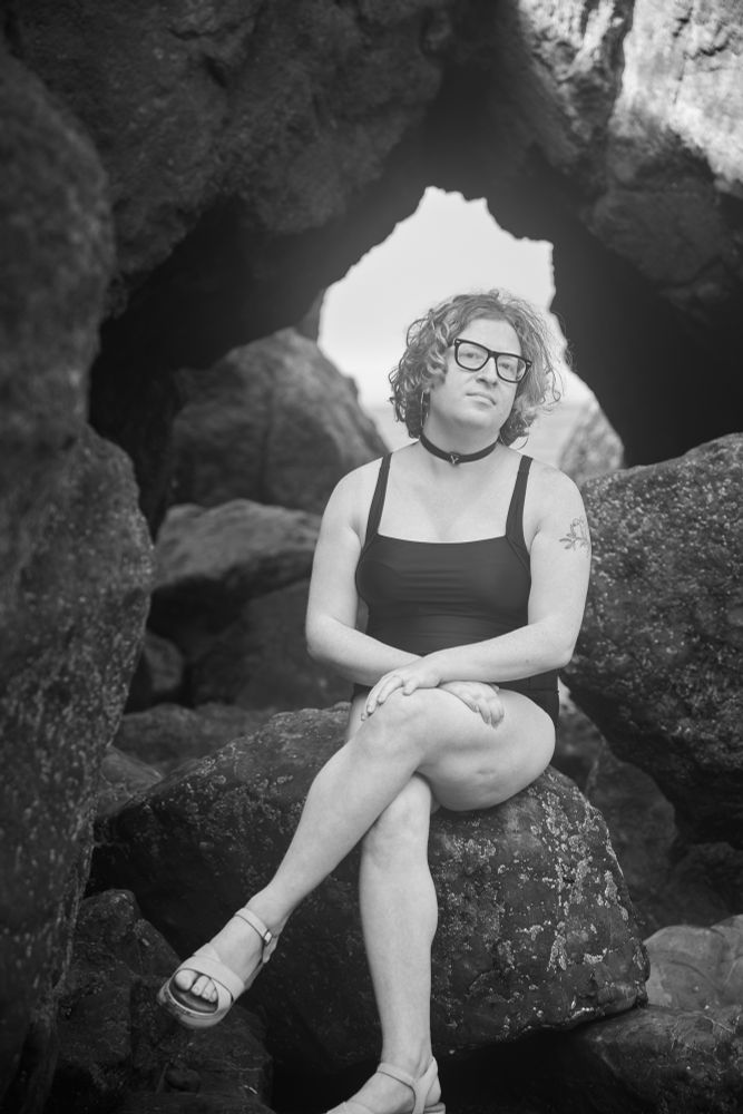 yael, in a black one piece bathing suit and white sandals, wearing a collar with a Y on it and large hoop earrings, sits on a rock in front of a rock arch in front of the ocean. the photo is black and white