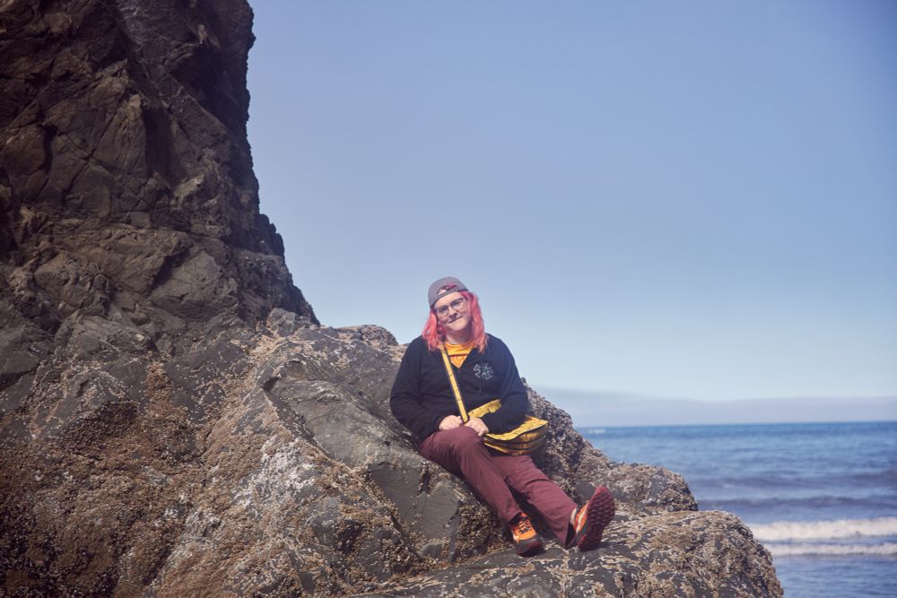 Noëlle sitting on a large rock in front of the ocean. e wears a union hoodie over a yellow shirt, with a yellow bag. e has a backwards baseball cap. e’s very cute