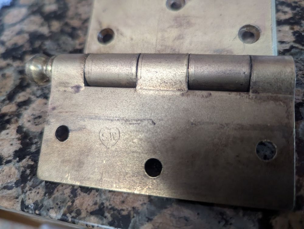 photo of slightly tarished brass hinge. A heart with the initials "S.W." inside is visible. 