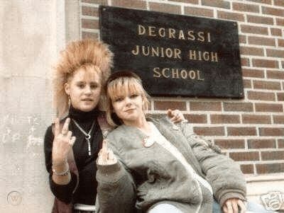 Two teen girls in front of a school sign that reads, "Degrassi Junior High School." One with spikey blonde hair has her fingers in a V, the other with blonde hair with bangs has her middle finger up. Both are dressed in a 1990s punk/grunge style 