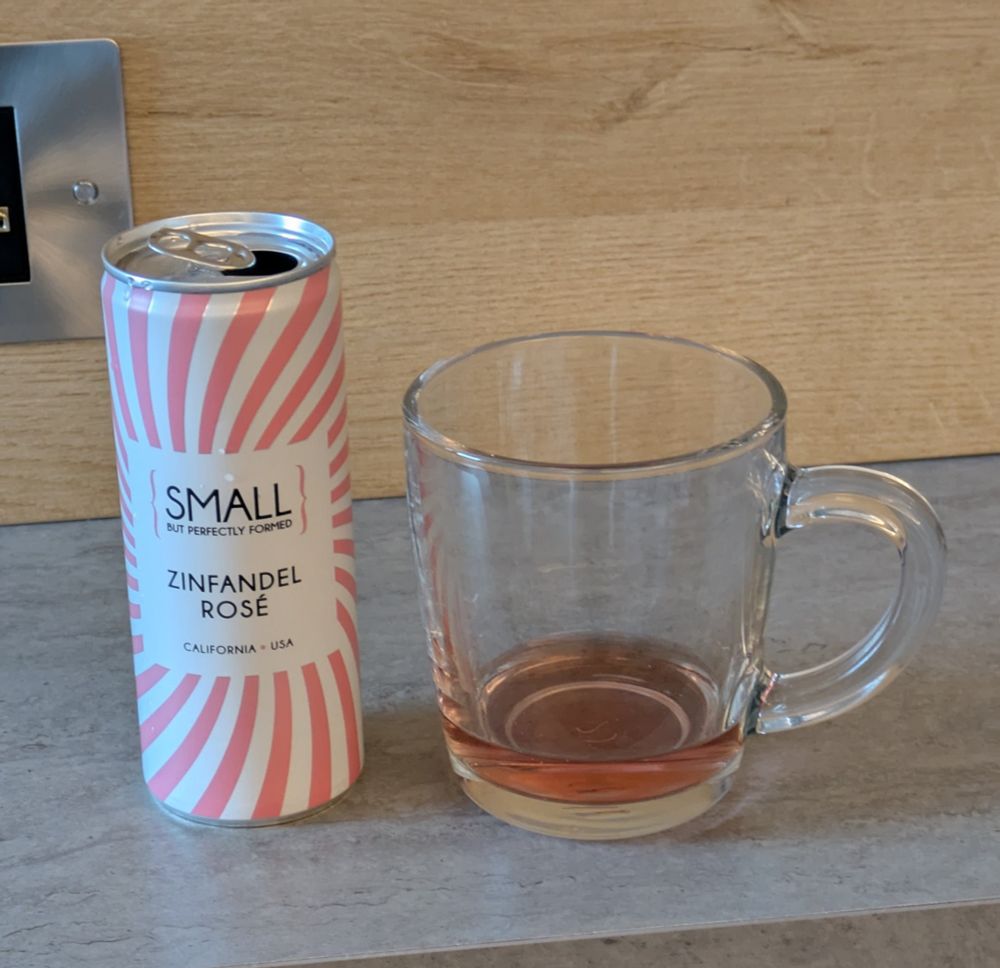 A pink and white striped can of zinfandel rose wine next to a glass mug with some pink coloured wine in it