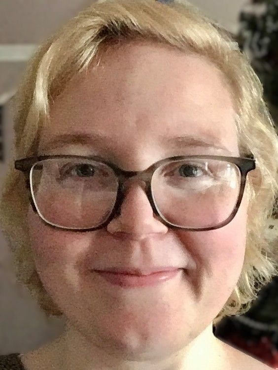 Photograph of Sarah Wride: short blonde hair, big glasses, smiling at the camera!