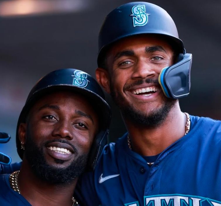 Randy Arozarena and Julio Rodriguez wearing helmets in the dugout, arms around each other smiling at the camera