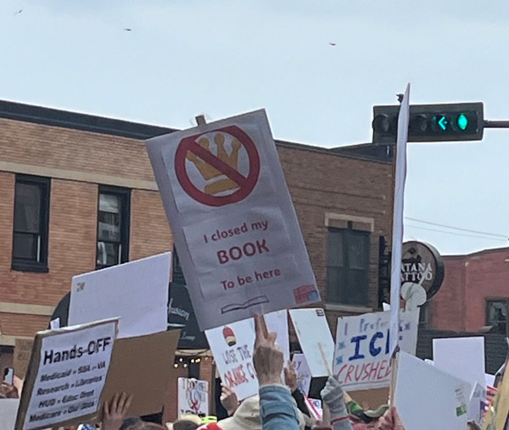 A sign with a crossed out crown that says, “I closed my book to be here.”