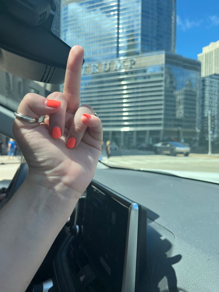 A picture of my hand from the passenger seat of my car. I have orange fingernails and a silver thumb ring. I’m flipping off trump tower and there is a cybertruck on the road in front of it. 