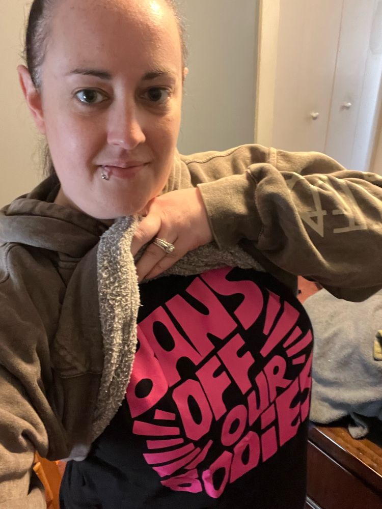 A selfie where I am wearing a hoodie and I’m lifting it up to show a t shirt underneath that says “Bans off our bodies” 