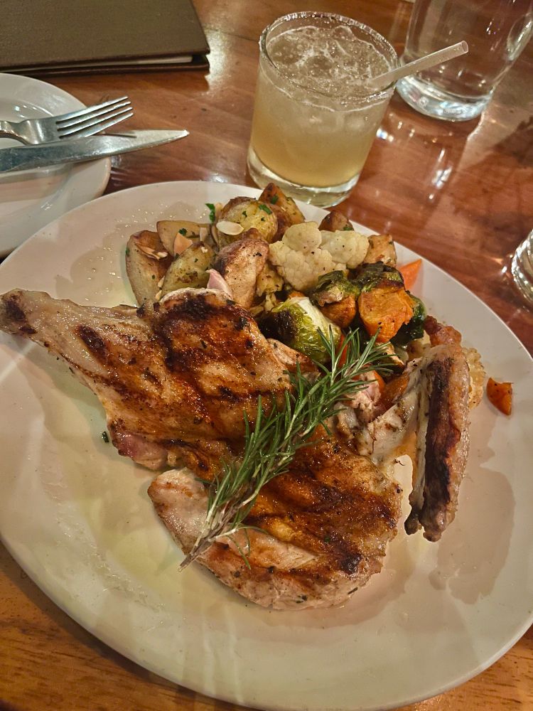 Picture of roasted chicken with an herb garnish on top. There is two sides: roasted and seasoned potatoes with almond slices. The other is cooked cauliflower, carrots, yams, and Brussels sprouts. 