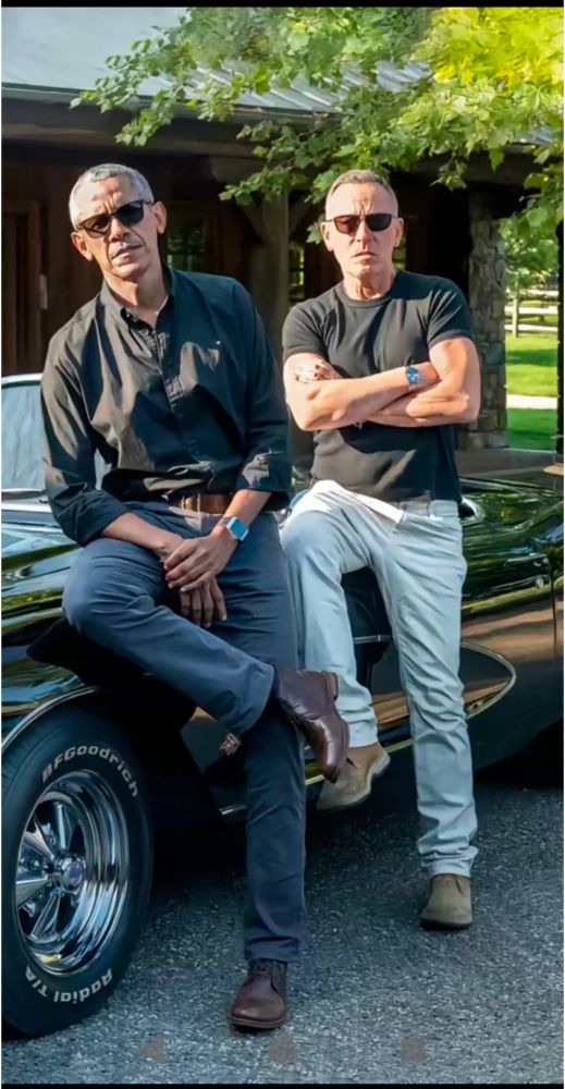Barack Obama and Bruce Springsteen lean against a car. Both wear sunglasses.