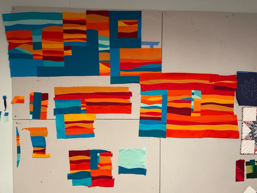 Pieces of a quilt in progress consisting of wavy strips of different shades of orange interspersed with solid blocks of aqua and wavy strips of different shades of aqua, meant to invoke the sun setting over the Pacific Ocean.