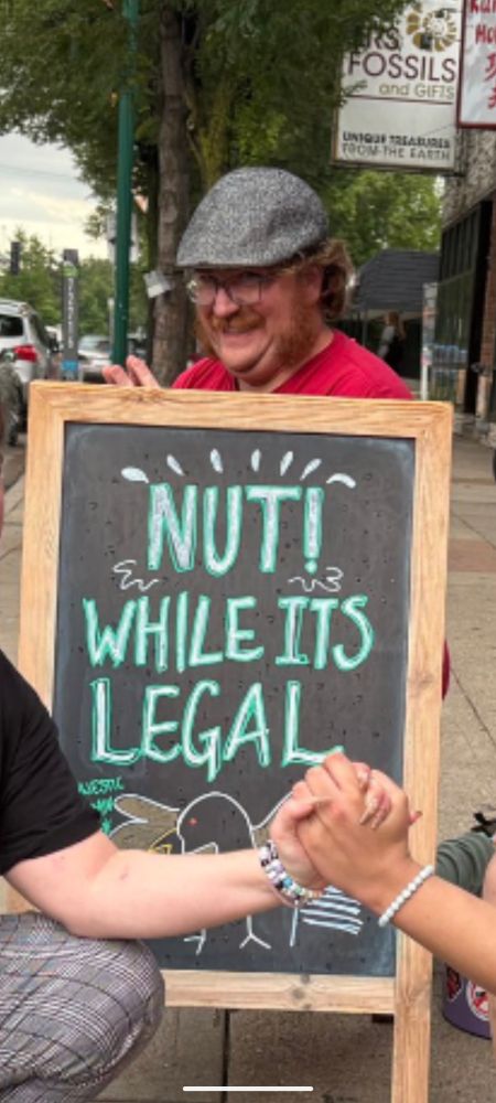 A zoomed in photo of a man making the worst face behind a chalkboard saying "NUT! WHILE IT'S LEGAL"