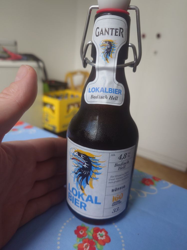 Eine Flasche Ganter Badisch Hell und die Hand des Users auf einem Küchentisch mit einer blauen Tischdecke mit Bildern von roten Rosen drauf. Im Hintergrund ein Kasten Mate.