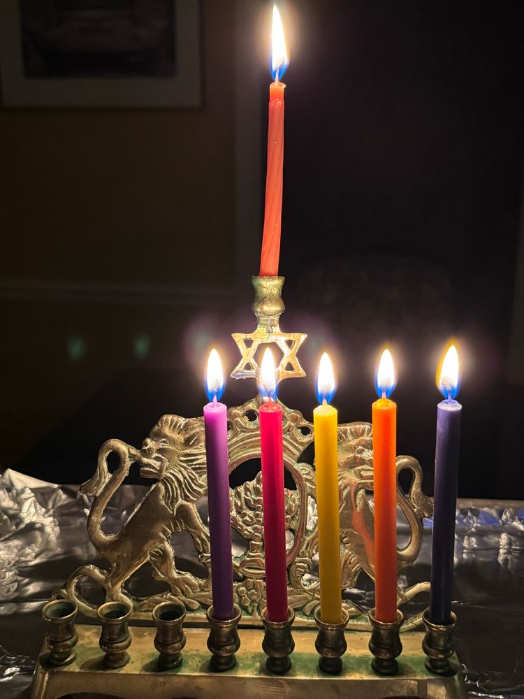 Antique brass Lion of Judah Chanukah menora (chanukiyah) with five candles lit
In front and the shammes (helper) candle at the top. It’s atop a table in a dark room.