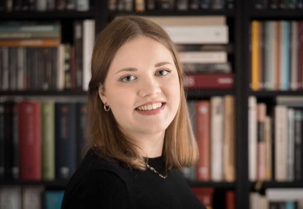 Portrait of Julia Płaczkiewicz. She is a white woman, posed standing up, smiling on a background of neatly stacked bookshelves. She is wearing a Black shirt. He hair is dark blonde, shoulder-length.