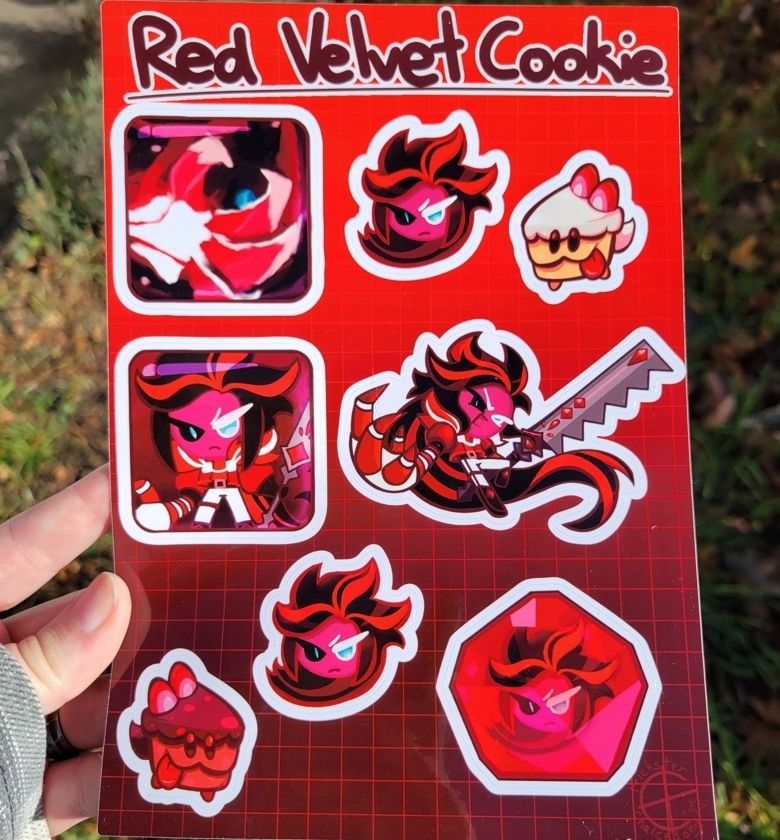 Image of a hand holding a red glossy sticker sheet with a grass background. Labeled "Red Velvet Cookie", it has stickers with his skill icons, headshot, gacha pose, soulstone, and cake hounds.