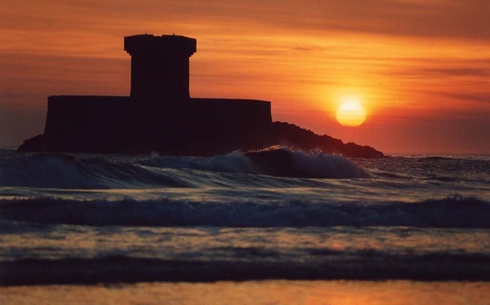 Sun sets behind a tower surrounded by sea. Waves break on the island and roll in towards the camera. 