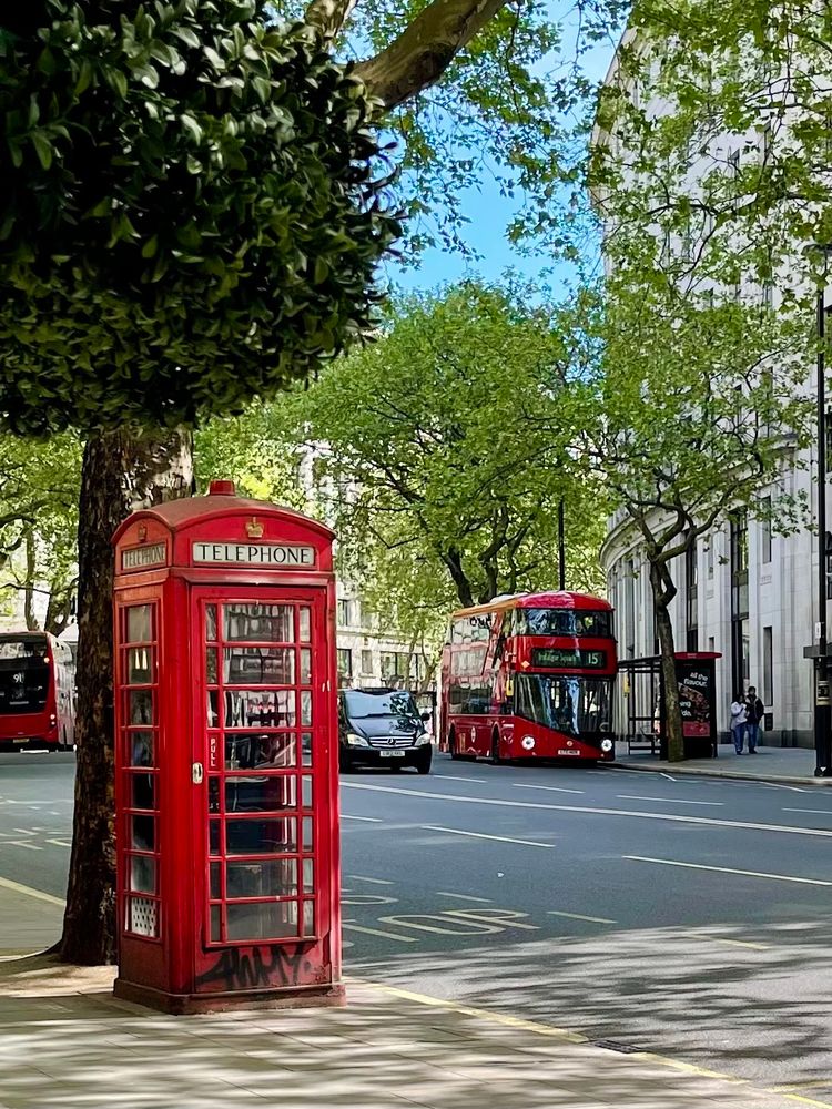 A close-up of a classic red British telephone box stands on the left side of a busy London street, partially obscured by the lush green leaves of a tree. In the background, red double-decker buses drive down the street, and a black car sits in traffic. The road is lined with sidewalks and a stately white stone building. The sky is clear and blue. 