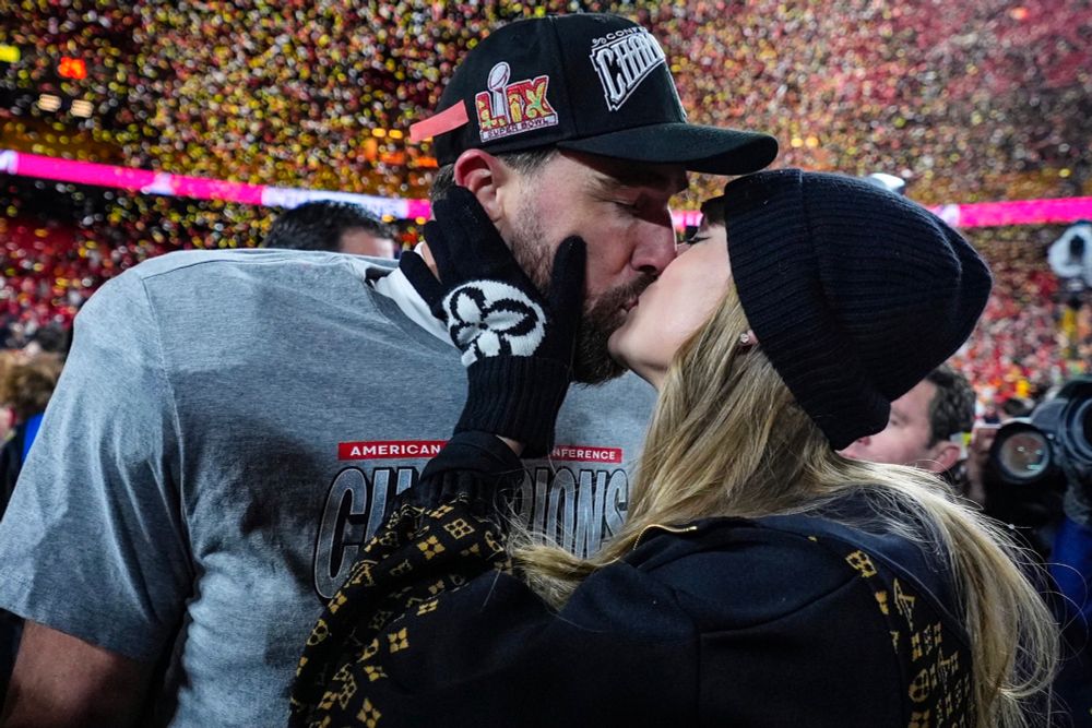 Taylor Swift and Travis Kelce celebrating Kansas City Chiefs’ AFC Championship win. They share a kiss as Taylor holds Travis’ face between her hands. An explosion of yellow and red confetti is seen in the background.