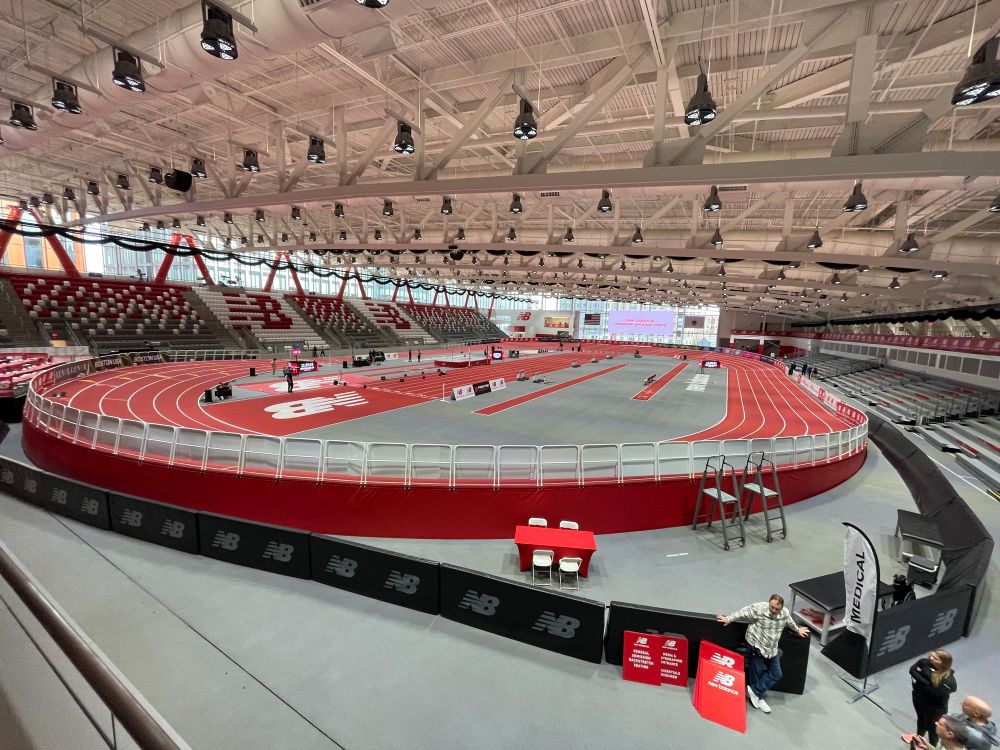 A track stadium at new balance, gray floors with a red track. A large screen hovers above displaying “New Balance Indoor Grand Prix”