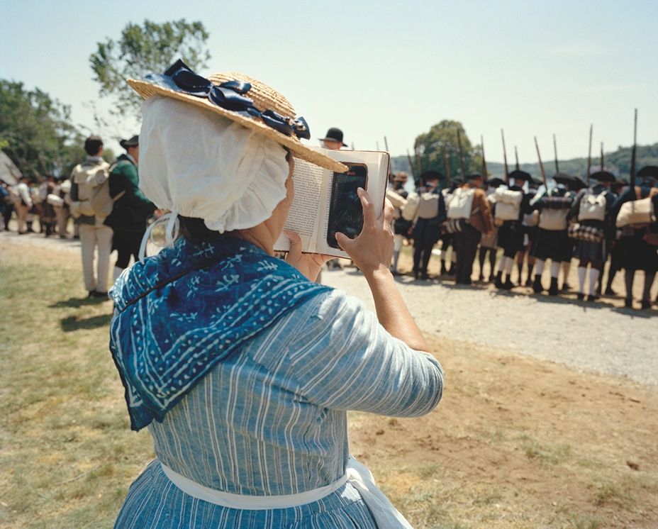 US Civil War reenactment, lady in blue dress and straw hat/bonnet thing has a phone hidden inside a hollowed out book