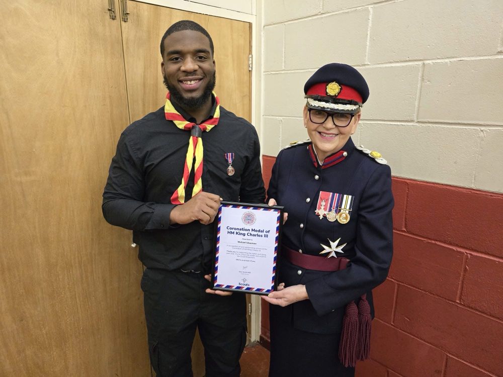 A person wearing a Scout scarf and a person in The uniform of a Deputy Lieutenant presenting a Coronation Medal.
