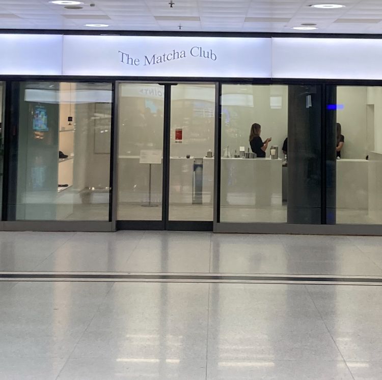 Frontansicht des Geschäfts “The Matcha Club”. Das Ladenlokal hat eine große Glasfront, durch die man eine Mitarbeiterin hinter einer weißen Theke erkennen kann. Das Schild über dem Eingang ist beleuchtet. Das Geschäft befindet sich in einem Gebäude mit hellgrauen Bodenfliesen, vermutlich einem Flughafen oder Einkaufszentrum.