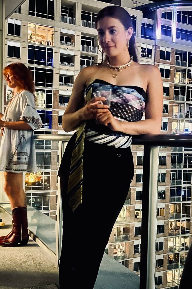 A girl wearing a top made out of men’s ties and a black skirt stands at a balcony.
