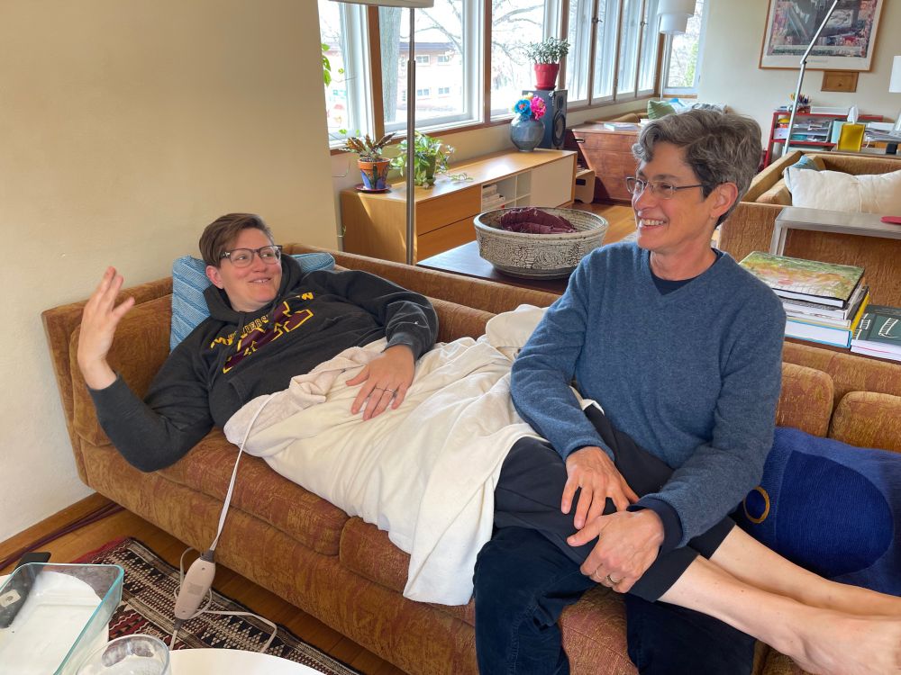 Two people sit on a couch in a cozy living room, smiling and talking. One is lying down under a blanket, and the other sits beside them with a hand on their legs.