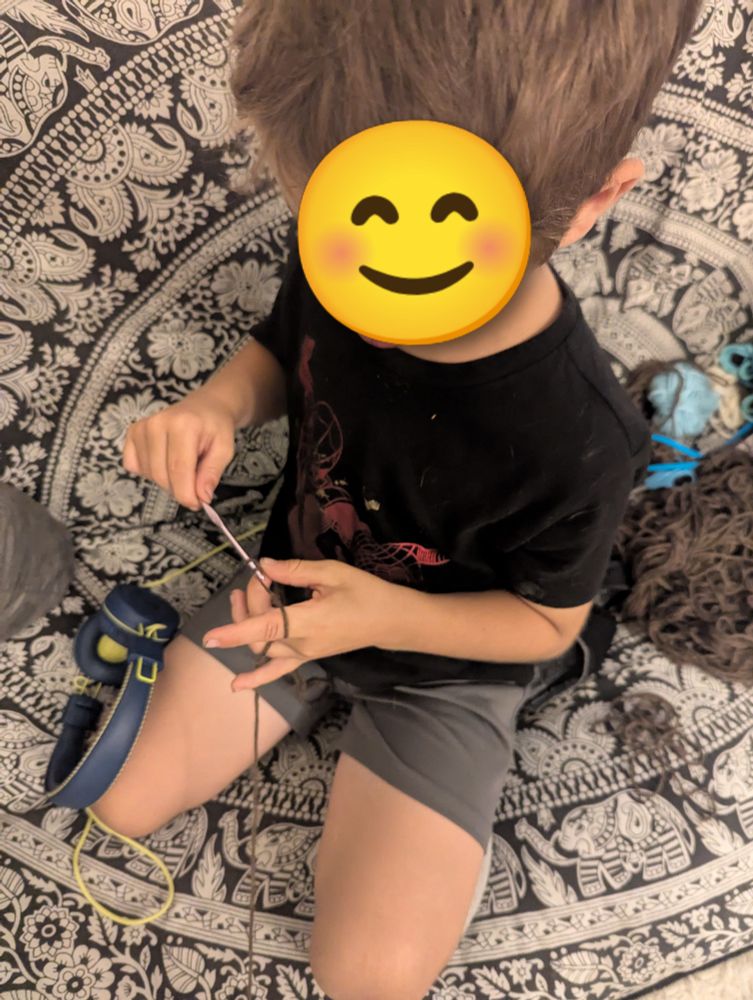 Six year old boy wearing Spider-Man T-shirt and grey shorts, on a couch, crocheting