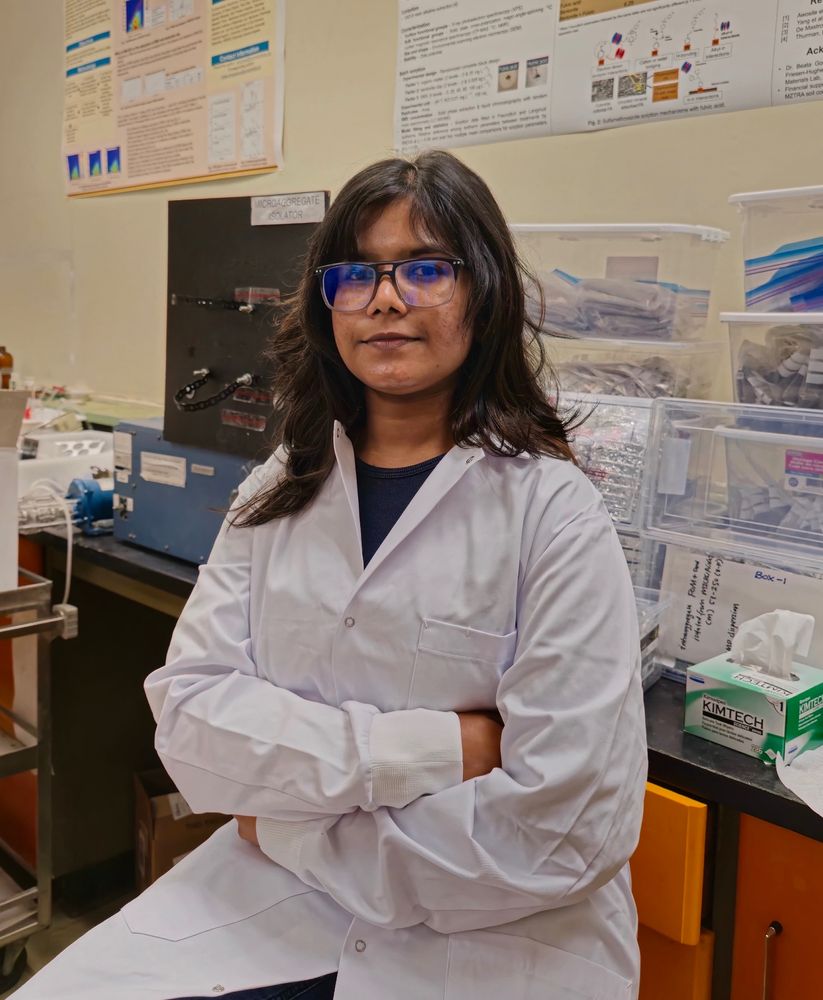 Photo of woman in lab coat sitting on a stool in front of a lab bench.