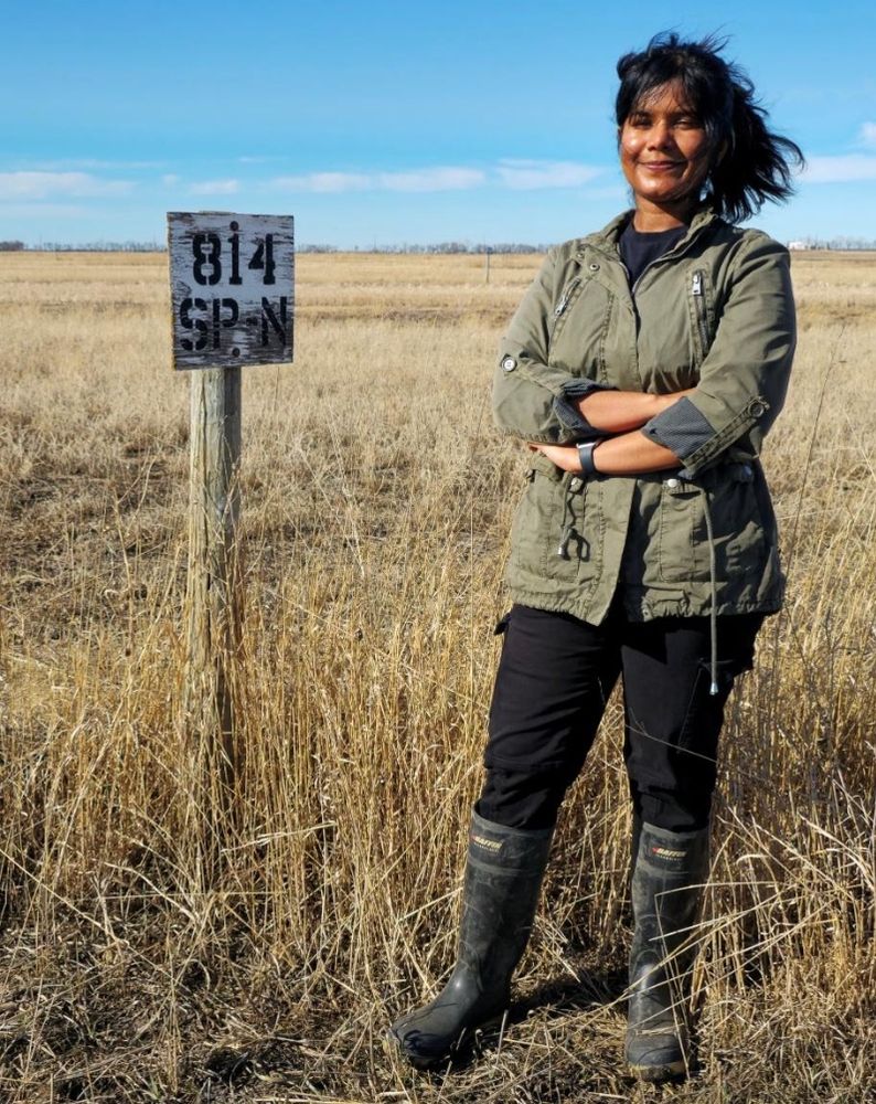 Person with shoulder length dark hair wearing a jacket, pants and boots, stands in a field. A blue sky is next to them.