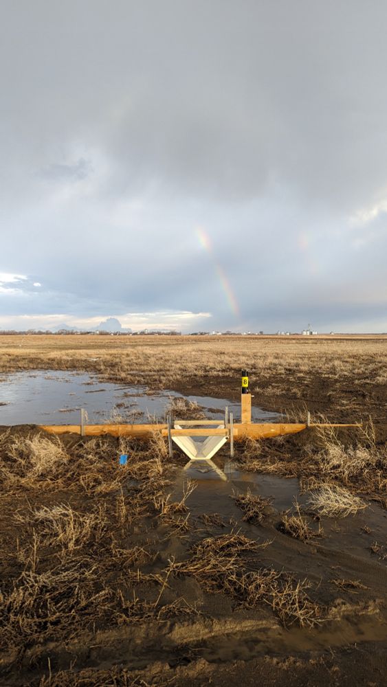 Equipment to measure water quality sits in a ditch along an agricultural field. In the background are clouds with a rainbow.