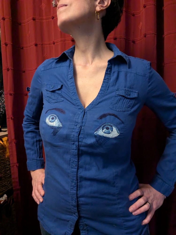 Une chemise bleue avec 2 grands yeux brodés en monochromie bleue au niveau de la poitrine, de part et d'autre des boutons.