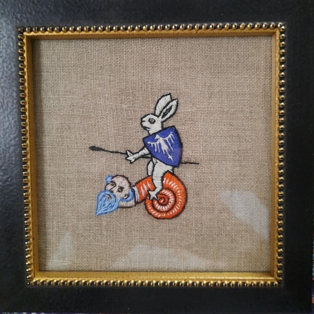 Broderie encadrée représentant un lapin avec une lance à cheval sur un homme escargot 
