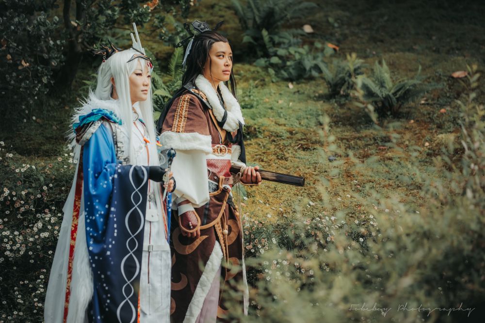 A photo of two people cosplaying Lin Xueya and Shang Buhuan from Thunderbolt Fantasy. They are posed as though taking a stroll through a blooming meadow.