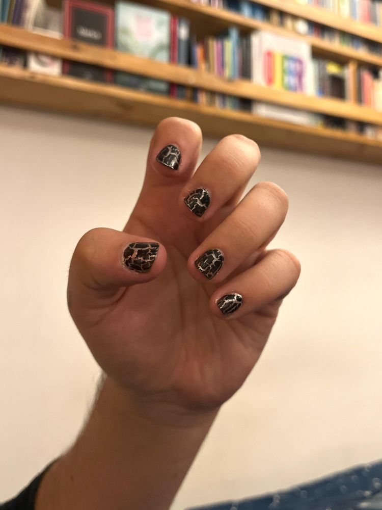 Mano con uñas pintadas de negro con grietas doradas, de fondo un librero con hermosos libros lgbtiqa+