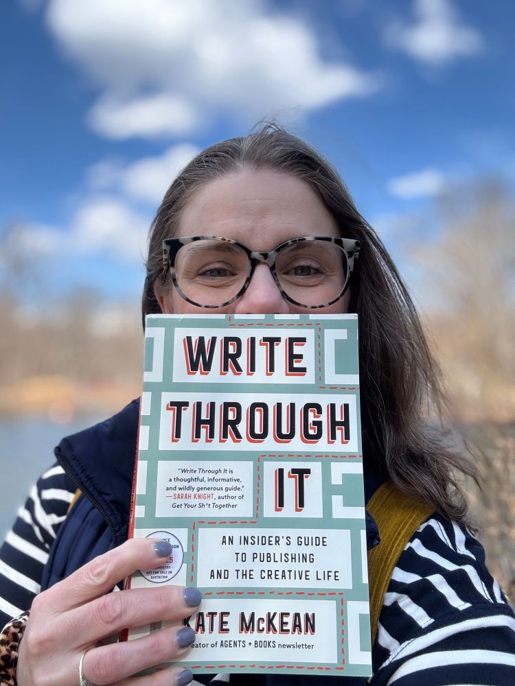 A white woman in glasses and a blue and white striped shirt standing outside on a sunny day holding a book in front of her face. The book’s title is Write Through It: and insider’s guide to publishing and the creative life by Kate McKean.