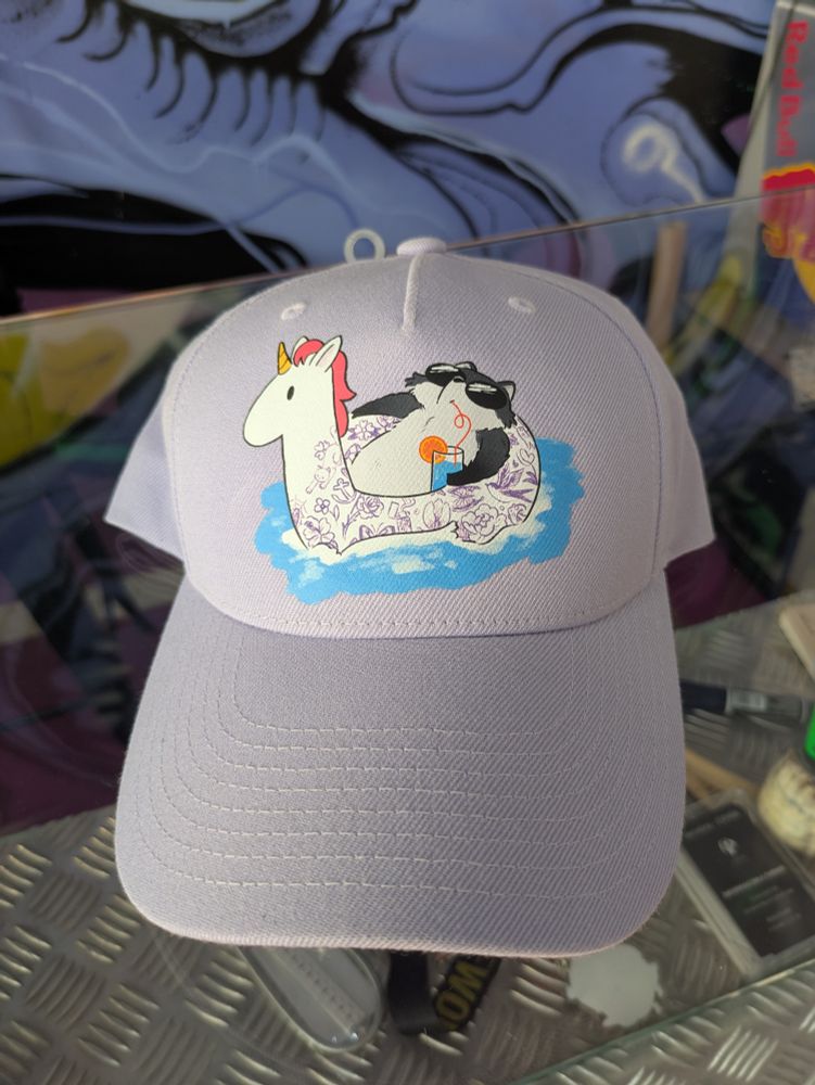 Gorra cerrada de color lila claro. En el centro de la frente, tiene la ilustración de un mapache con gafas de sol, tomándose un cóctel azul en un flotador de unicornio con el pelo rosa y tatuajes en lila.