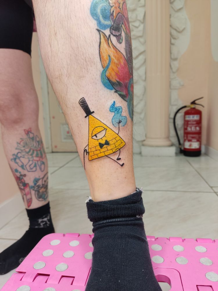 Foto de un tatuaje acabado de hacer en la parte inferior de la pierna, por encima del tobillo. El tatuaje es Bill, un personaje de la serie Gravity Falls. Bill es una pirámide 2D con brazos y piernas, un ojo en medio de la pirámide, una pajarita negra debajo del ojo y un sombrero de copa alto en la punta. Bill está en una posición desenfadada, con un bastón colgando del brazo que queda a la izquierda de la imagen, y una llama azul en la mano que queda a la derecha de la imagen. La pirámide es de un color amarillo compacto, mientras que el sombrero negro y la llama azul estan coloreados como si estuvieran garabateados con lápiz y sombreados por encima.
En el fondo de la imagen a la derecha, desenfocado, hay un extintor, mientras que la otra pierna de la persona tatuada queda a la izquierda de la imagen.