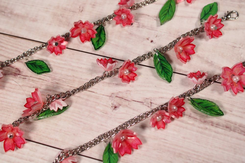 Three similar bracelets all laid out parallel to each other. Each bracelet is made up of a silver chain with small pink flowers and green leaves attached to it.