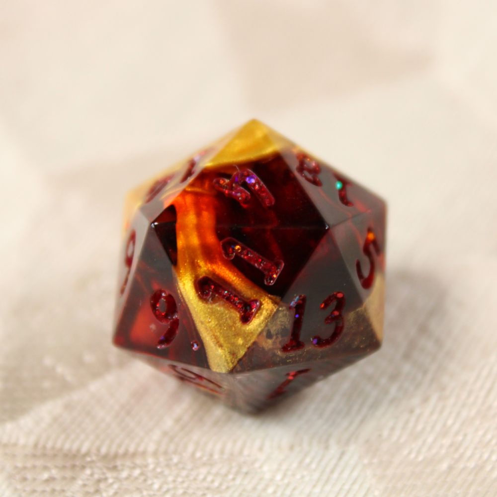 A close up of a single d20 die. The die is dark red and has shimmering drips of yellow-gold through it. The numbers are also dark red but contain glitter.