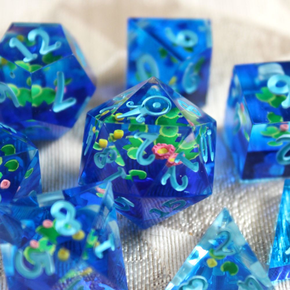 A close up of a d20 die surrounded by other dice in varying shapes. The d20 contains hand-painted lily pads and tiny polymer clay flowers floating over a layer of dark blue. The numbers are light blue.