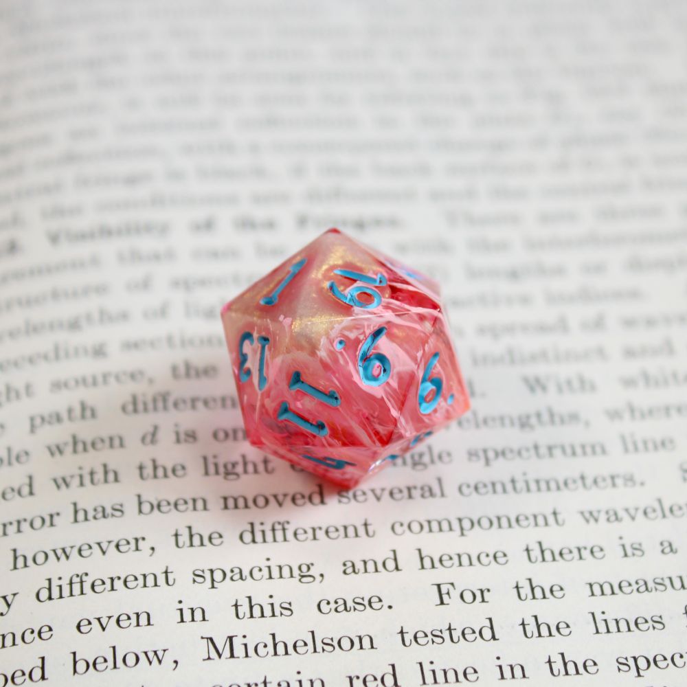 A single bright orange-pink d20 with bright blue numbers. The die is very peachy in color from this angle, and has a pale gold shimmer at one end. The die is resting on the page of a book.